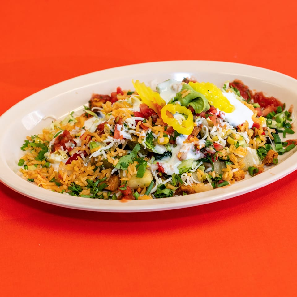 Loaded Mexican-style rice bowl topped with lettuce, cheese, salsa, guacamole, sour cream, peppers, corn, and cilantro on a white platter.