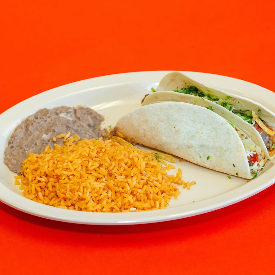 Plate of three soft tacos with lettuce, cheese, and vegetables, served with Mexican rice and refried beans on a red tablecloth.