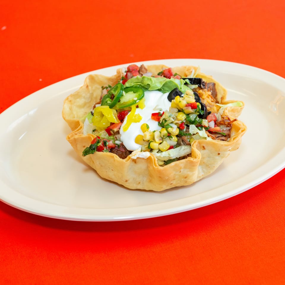 Crispy tortilla bowl filled with seasoned meat, lettuce, sour cream, black olives, peppers, tomatoes, and corn salsa on a white plate.