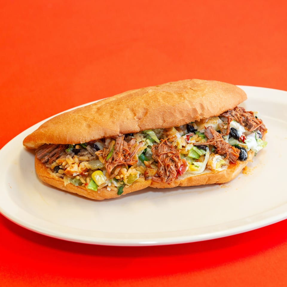 Loaded toasted sandwich with shredded meat, beans, rice, lettuce, vegetables, and creamy sauce on a white plate.