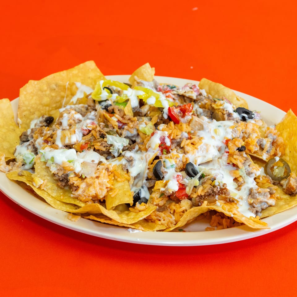 Loaded plate of nachos topped with seasoned meat, rice, beans, vegetables, black olives, jalapeños, and creamy cheese sauce.